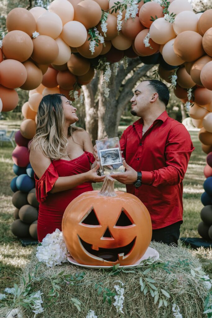 Couple with a sonogram at an outdoor baby shower with a jack-o-lantern and balloon arch